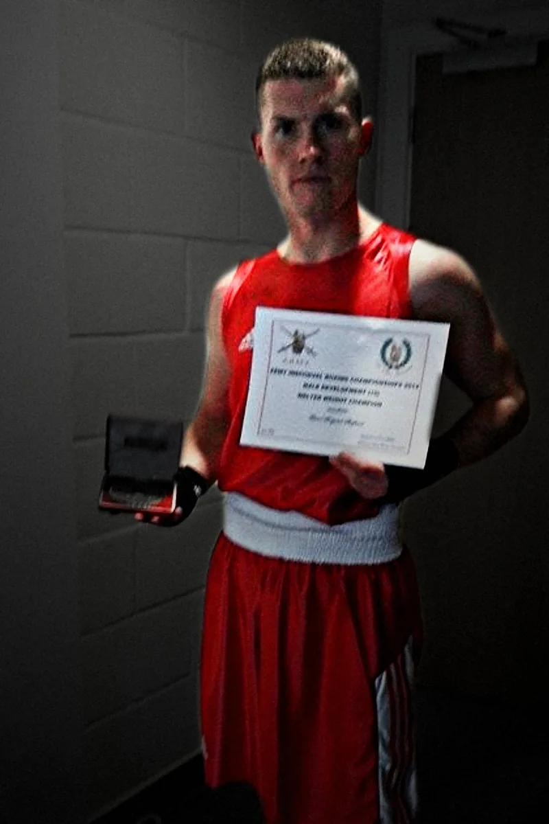 A boxer in Army red holding his medal and his Army boxing championships certificate