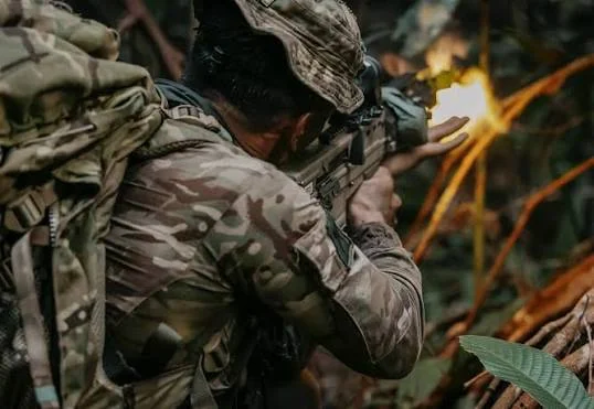 A soldier in camouflage firing a rifle in thick jungle